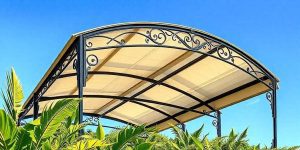 A decorative metal gazebo with a cream-colored canopy roof stands among green leafy plants, set against a clear blue sky.