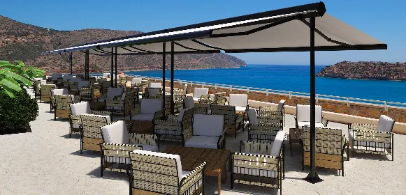 Outdoor terrace with cushioned wicker chairs and tables under a large awning, overlooking a blue sea and rocky hills on a sunny day.