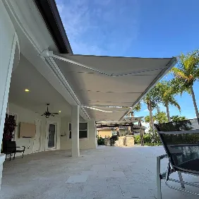 A modern patio with a large retractable awning extended, providing shade over a tiled outdoor seating area. Palm trees and a clear blue sky are visible in the background.