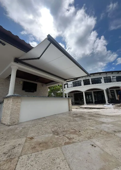 A modern outdoor patio with a stone bar, white retractable awning, and tiled flooring. In the background, there is a large, two-story white building with columns under a partly cloudy sky.