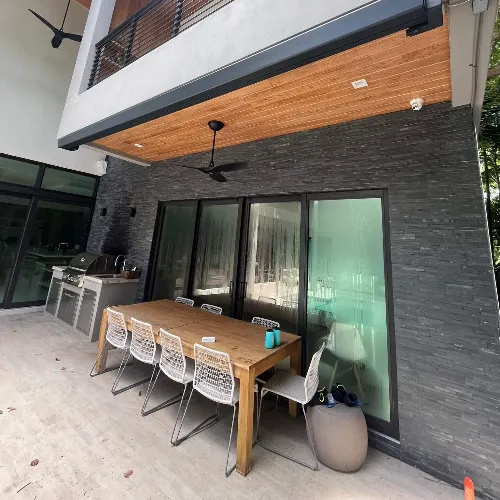 Modern outdoor patio with a wooden dining table, six white woven chairs, a built-in grill, and large glass doors under a covered ceiling with a fan, next to a dark stone accent wall.
