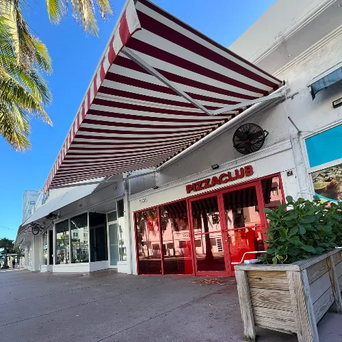A sidewalk view of a white building with red-framed windows and a red-and-white striped awning reading &quot;Pizza Club.&quot; Palm tree leaves and a planter with greenery are visible in the foreground.