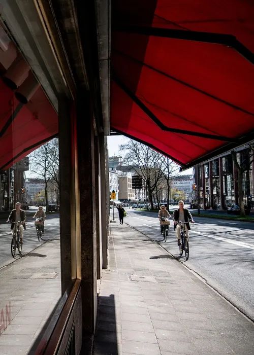 Two people ride bicycles down a city street under a large red awning. Their reflections appear in a shop window, and trees and buildings line the background on a sunny day.