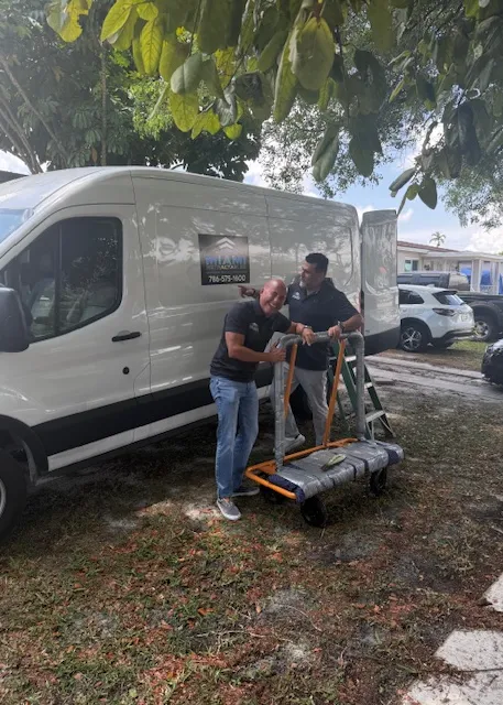 Two men stand beside a white van under trees, smiling as they move a large wrapped item on a hand truck. The van has a business sign and a ladder is visible near the back of the vehicle.