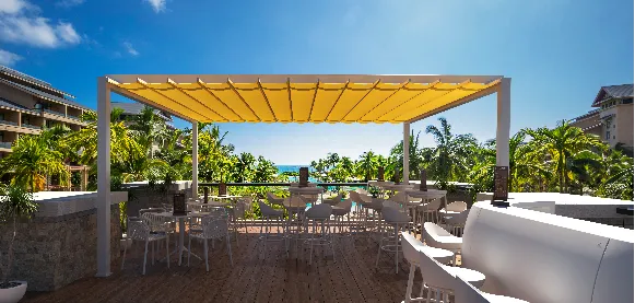 Outdoor terrace with white tables and chairs under a yellow pergola, surrounded by palm trees, overlooking the ocean on a sunny day.