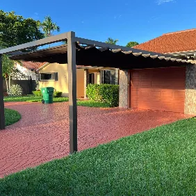 A suburban house with a brown garage door, a red stamped concrete driveway, a pergola-style carport, green grass, and a green trash bin near the garage. Trees and other houses are visible in the background.
