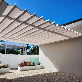 A modern white fabric pergola provides shade over a gravel patio with raised white planters filled with green plants and flowers, against a backdrop of blue sky and a white fence.