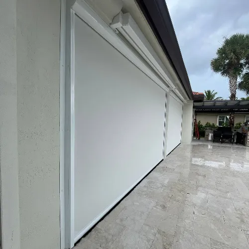 Large white storm shutters cover the windows and door of a building with a tiled patio. A palm tree and outdoor furniture are visible in the background under a cloudy sky.