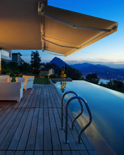 Modern patio with a wooden deck, white outdoor furniture, and an infinity pool overlooking a scenic mountain and lake view at sunset. A large retractable awning is extended above the seating area.