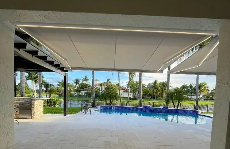 A view from a covered patio looking out at a backyard pool with blue tiles, surrounded by palm trees and a grassy lawn, with houses visible in the background.