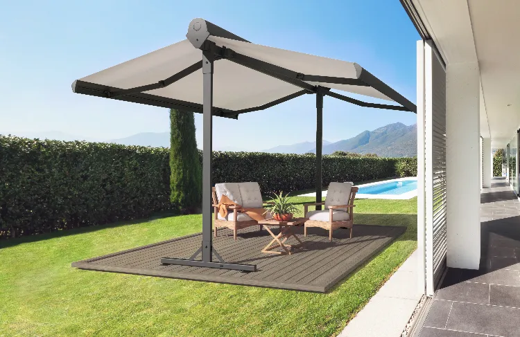 A patio with two cushioned chairs, a table, and plants sits under a large dual-canopy umbrella beside a pool, bordered by a tall hedge and mountains in the background on a sunny day.