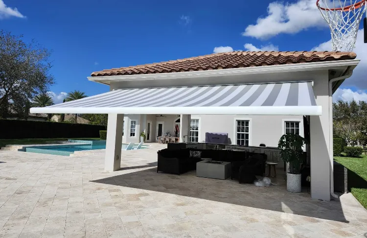 A modern outdoor patio with beige tiles features a retractable striped awning, providing shade over a seating area. A swimming pool and green lawn are visible under a clear blue sky.
