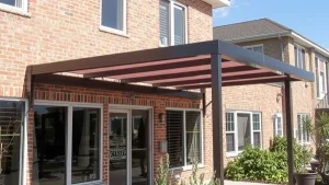 A modern metal pergola with open beams is attached to the back of a brick house, covering a patio area with large glass doors and windows. Green plants and neighboring houses are visible in the background.