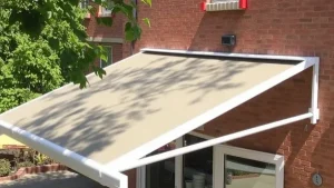 A beige retractable awning extends over a patio door on a brick house, casting shade. Green tree branches with leaves are visible in the foreground, creating shadows on the awning.