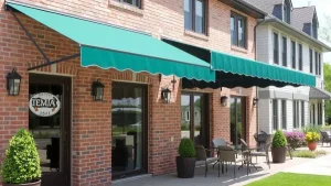 A brick building with green and white striped awnings over patio areas, featuring black lantern lights, potted plants, outdoor tables and chairs, and large windows.