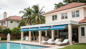 A modern house with a red-tiled roof features a covered patio with blue awnings, four white lounge chairs, and a swimming pool in the foreground. Palm trees and greenery surround the backyard.