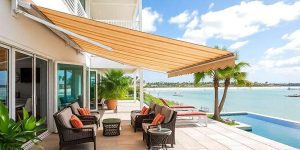 A patio with wicker chairs and cushions under a striped retractable awning overlooks a pool and a scenic waterfront with boats and palm trees on a sunny day.