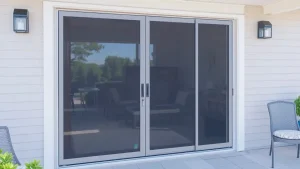 A modern glass sliding patio door with a screen, set in a light-colored exterior wall. Outdoor chairs are visible on the tiled patio, and the room inside is partially visible through the glass.