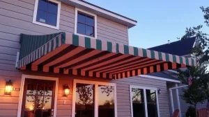 A house with beige siding features a large, green-and-white striped retractable awning over a patio area, with outdoor lights on either side of the glass doors at sunset.